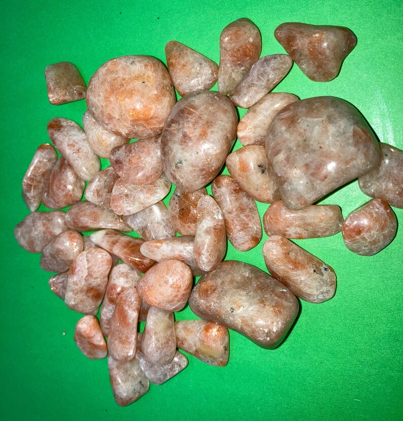 May include: A collection of tumbled sunstone crystals in various sizes and shapes. The stones are a light orange color with a shimmery, iridescent finish.