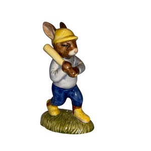 May include: A ceramic figurine of a rabbit dressed in a baseball uniform, holding a bat. The rabbit wears a yellow cap, blue trousers, and yellow shoes. It stands on a patch of green grass.