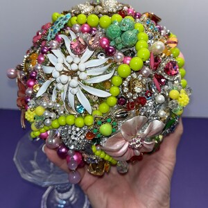 May include: A round bouquet made of various vintage brooches, rhinestones, and beads. The bouquet is predominantly green, pink, and white, with a large white flower in the center.