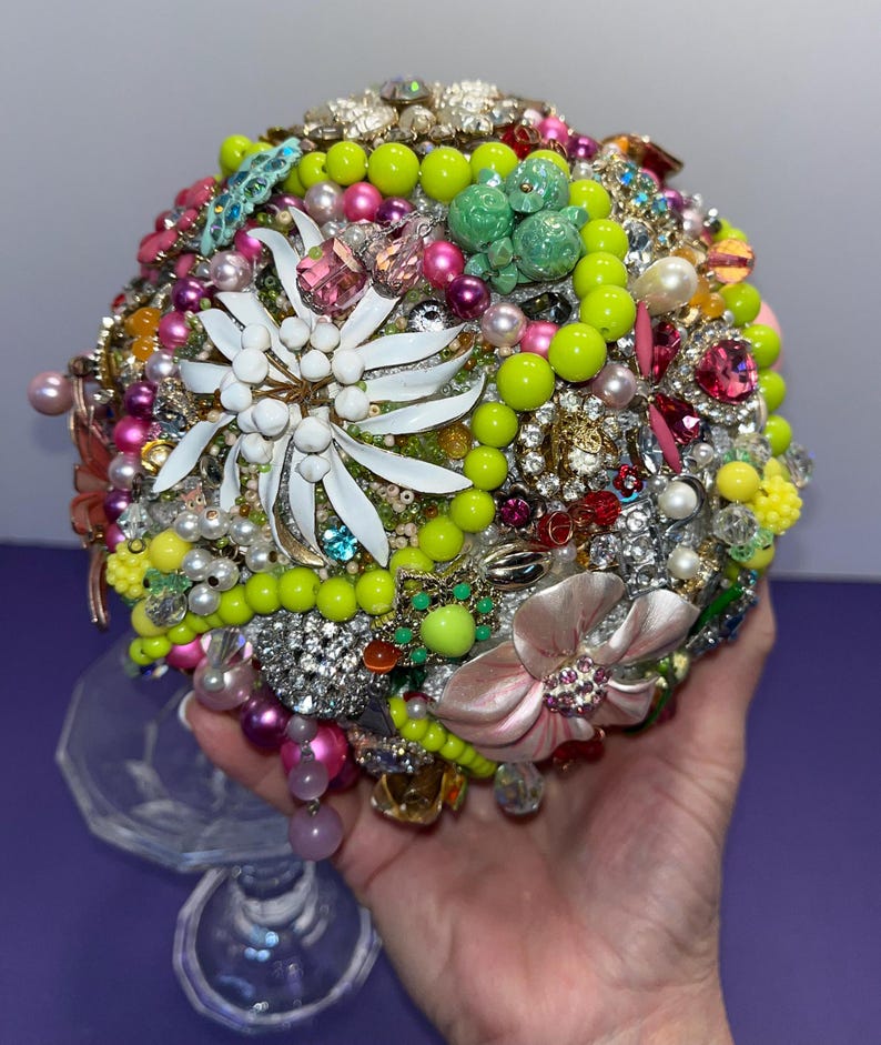 May include: A round bouquet made of various vintage brooches, rhinestones, and beads. The bouquet is predominantly green, pink, and white, with a large white flower in the center.