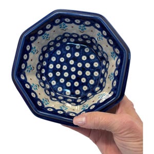 May include: An octagonal ceramic bowl with a blue and white floral pattern. The bowl has a dark blue rim and a detailed interior design featuring small white dots and floral accents. The bowl is held by a hand.