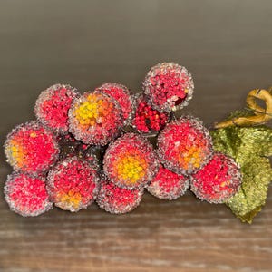Pode incluir: Um cacho de uvas artificiais com bagas vermelhas e amarelas, cobertas com uma substância brilhante. As uvas estão presas a uma folha verde e a um caule dourado, sobre um fundo de madeira.