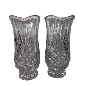 May include: Two clear glass vases with a detailed, cut-glass design. The vases have a flared top and a diamond-patterned base. The clear glass reflects light, creating a sparkling effect. The vases are identical in shape and size.