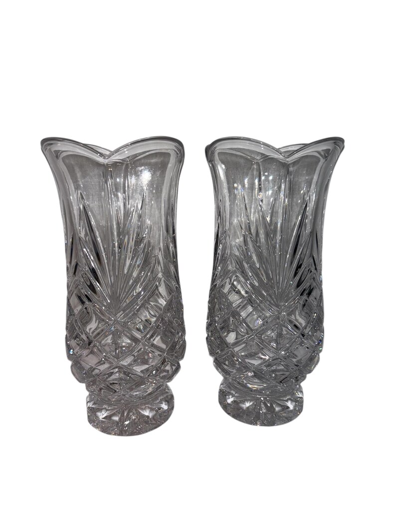 May include: Two clear glass vases with a detailed, cut-glass design. The vases have a flared top and a diamond-patterned base. The clear glass reflects light, creating a sparkling effect. The vases are identical in shape and size.