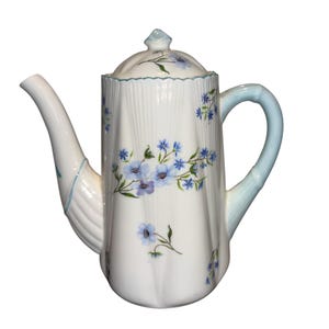 Puede incluir: Tetera de cerámica blanca con tapa, boquilla y asa. La tetera está decorada con motivos florales azules y un asa azul claro. El cuerpo tiene detalles acanalados verticales. La tapa tiene un pequeño adorno y un ribete verde.