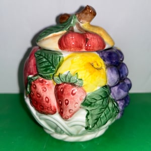 Könnte beinhalten: Eine Keramik-Zuckerdose in Form eines Obstkörbchens mit Deckel. Die Dose ist mit verschiedenen Früchten verziert, darunter Erdbeeren, Trauben, Kirschen und eine Birne. Die Dose ist weiß mit grünen Blättern und einem braunen Stiel.