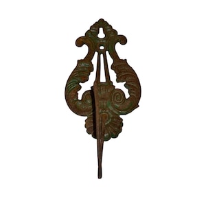 Puede incluir: Gancho de metal de color bronce antiguo con un diseño ornamental en forma de voluta. El gancho tiene una parte inferior larga y puntiaguda y una sección superior detallada con patrones arremolinados y un toque de pátina verde.