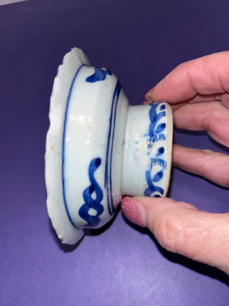 May include: A small, white ceramic bowl with a blue and white floral design. The bowl has a scalloped edge and a raised base.