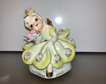 Może przedstawiać: Vintage ceramiczna figurka aniołka w żółtej sukience, trzymającego kwiaty. Figurka ma złote akcenty, aureolę i małe skrzydła. Na podstawie napis "SATURDAY CHILD".
