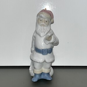 May include: A white porcelain Santa Claus figurine with a pink hat, blue boots, and a white beard. He is holding a bag of gifts in his left hand.