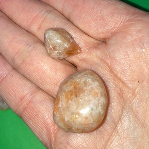 May include: Two sunstone tumbled stones, one is a rounded shape and the other is a triangular shape. Both stones are a light orange color with a shimmery, iridescent quality.