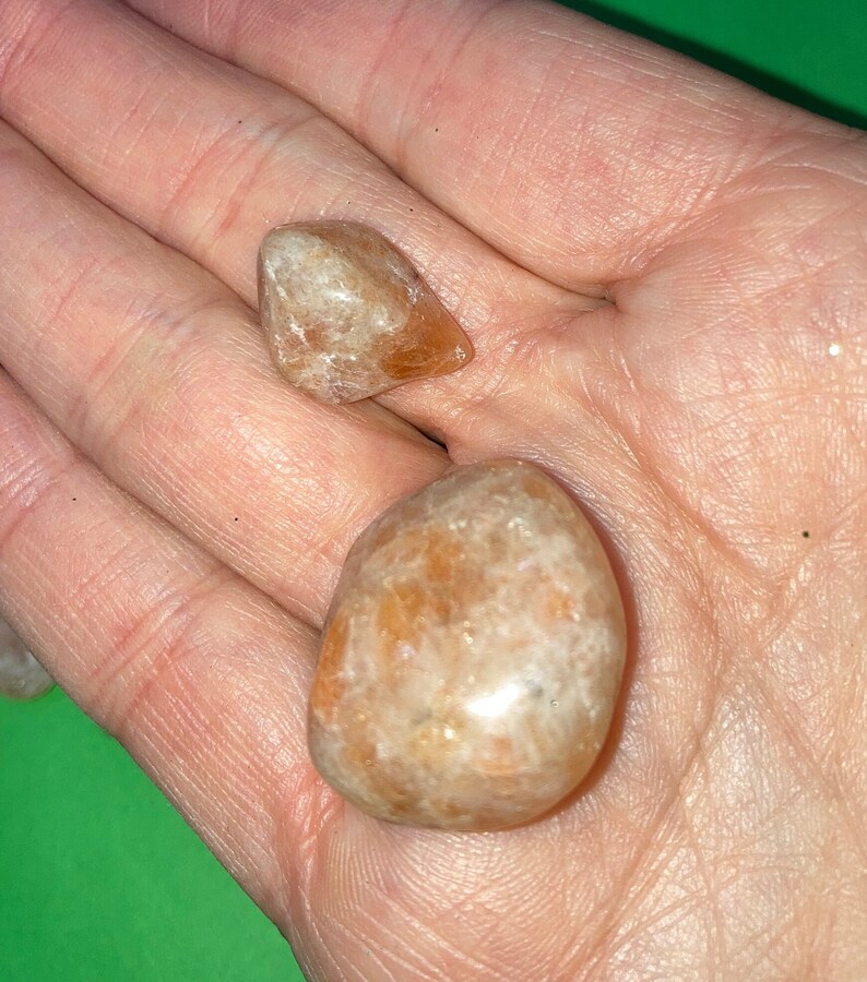 May include: Two sunstone tumbled stones, one is a rounded shape and the other is a triangular shape. Both stones are a light orange color with a shimmery, iridescent quality.