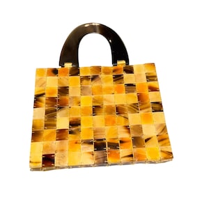May include: A square handbag with a woven pattern in shades of yellow, brown, and cream. The bag has a dark brown, arched handle. The woven design creates a textured appearance, and the bag is set against a white background.