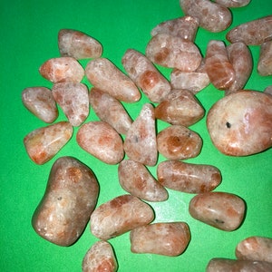 May include: A collection of tumbled sunstone crystals in various sizes and shapes. The stones are a light orange color with a shimmery, iridescent finish.