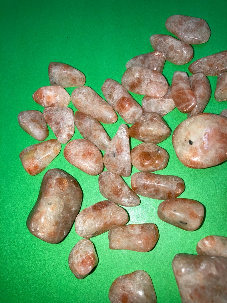 May include: A collection of tumbled sunstone crystals in various sizes and shapes. The stones are a light orange color with a shimmery, iridescent finish.