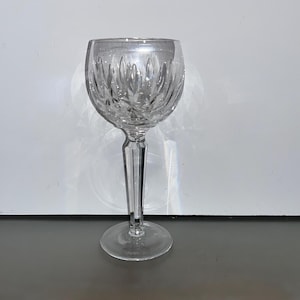 May include: A clear crystal wine glass with a detailed, etched pattern on the bowl. The glass has a long, slender stem and a round base. The bowl features a repeating leaf-like design.