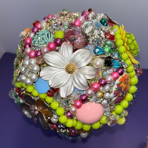 May include: A colorful, round bouquet made of various beads, rhinestones, and flowers. The bouquet is primarily green, pink, and white, with a large white flower in the center.