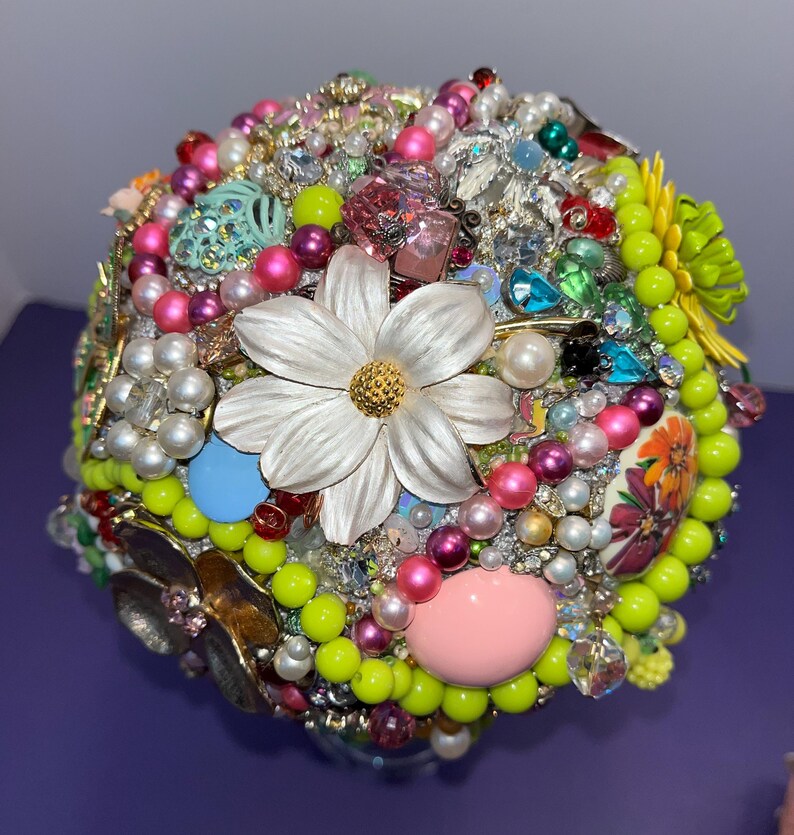 May include: A colorful, round bouquet made of various beads, rhinestones, and flowers. The bouquet is primarily green, pink, and white, with a large white flower in the center.