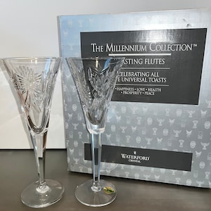 Op de afbeelding: Twee champagnefluiten van Waterford-kristal met een vlinderontwerp. De fluiten maken deel uit van The Millennium Collection, ter ere van alle universele toasts. De doos heeft de tekst "The Millennium Collection™  Tasting Flutes  Celebrating All The Universal Toasts  Happiness Love Health Prosperity Peace  Waterford Crystal".