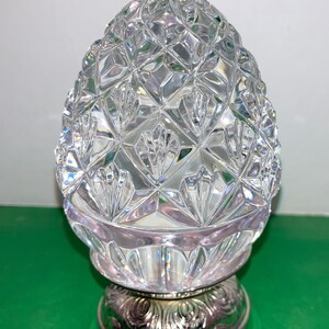 May include: A clear, cut-glass decorative object with a pineapple-like shape. The top portion features a diamond pattern, while the base is silver-colored with an ornate design. The object sits on a green surface.