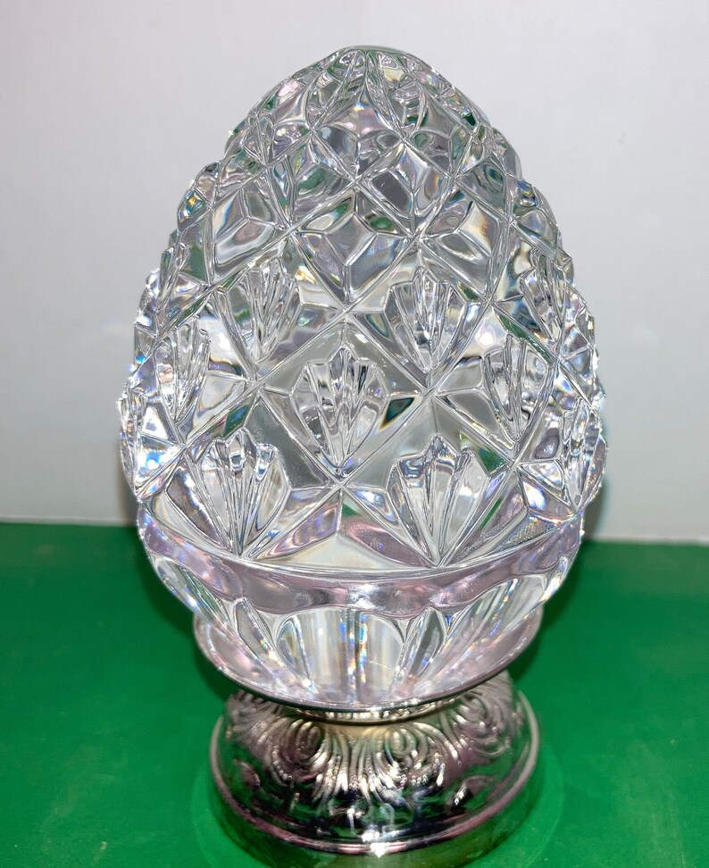May include: A clear, cut-glass decorative object with a pineapple-like shape. The top portion features a diamond pattern, while the base is silver-colored with an ornate design. The object sits on a green surface.