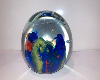 May include: A clear glass paperweight with blue, green, and orange swirls and bubbles inside.