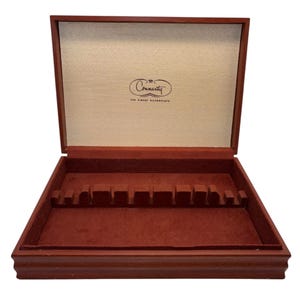 May include: Open wooden silverware storage box with a red velvet interior. The box has a hinged lid and a series of slots for holding silverware. The lid has the text "Community The Finest Silverplate".