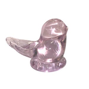 May include: A translucent, light purple glass bird figurine. The bird is perched on a circular base, with its wings slightly outstretched. The glass has a smooth, reflective surface, catching the light.