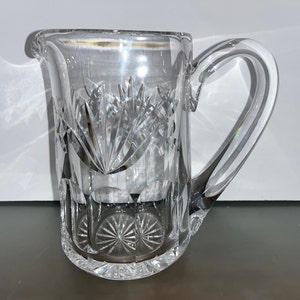 May include: A clear glass pitcher with a cut glass design. The pitcher has a handle and a round base.