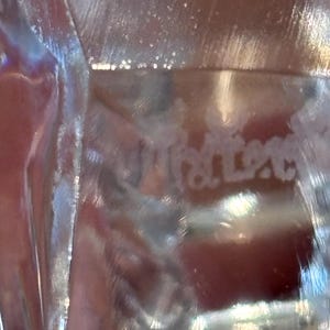 K&ouml;nnte beinhalten: Nahaufnahme eines transparenten Glasobjekts mit dem Wort "Kristal" in die Oberfl&auml;che ge&auml;tzt. Das Glas hat eine reflektierende Qualit&auml;t, wobei sich das Licht durch es hindurch bricht und einen schimmernden Effekt erzeugt. Der Hintergrund ist eine verschwommene Farbmischung.