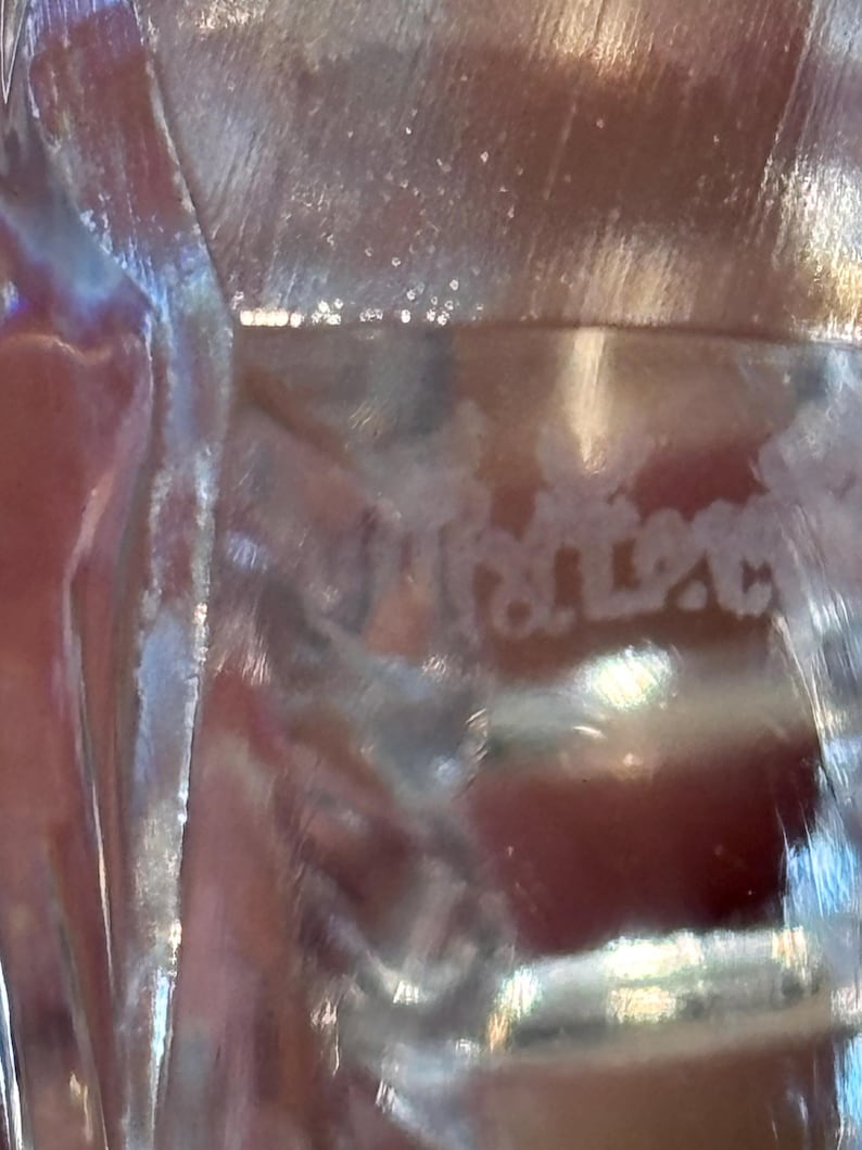K&ouml;nnte beinhalten: Nahaufnahme eines transparenten Glasobjekts mit dem Wort "Kristal" in die Oberfl&auml;che ge&auml;tzt. Das Glas hat eine reflektierende Qualit&auml;t, wobei sich das Licht durch es hindurch bricht und einen schimmernden Effekt erzeugt. Der Hintergrund ist eine verschwommene Farbmischung.