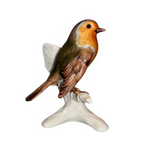May include: A ceramic figurine of a robin perched on a white branch. The bird has a vibrant orange breast, a brown back, and a dark beak. The figurine is set against a white background.