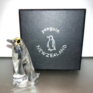 May include: A clear glass penguin figurine with a yellow eye, sitting on a black box with the text "penguin NEW ZEALAND".