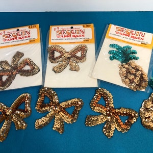 Può includere: Un set di sei applicazioni in paillettes dorate e turchesi. Le applicazioni hanno la forma di fiocchi e ananas. Le applicazioni sono confezionate su schede con il testo "Hand Embroidered Sequin Appliques for Sweaters, Jeans, Blouses, Dresses etc. $ 1,89".