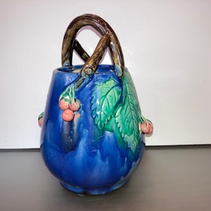 Puede incluir: Un jarrón de cerámica azul con un asa de rama y hojas verdes y bayas rosas. El jarrón está esmaltado con un patrón de remolino azul y blanco.