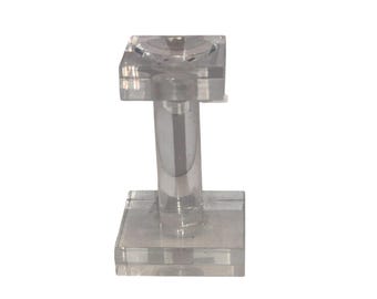May include: A clear, acrylic jewelry display stand. The stand has a square base, a cylindrical stem, and a square top with a small, recessed area. The design is simple and modern, suitable for showcasing rings or small items.