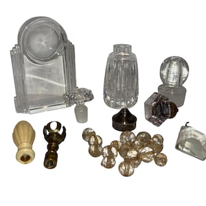May include: A collection of vintage glass perfume bottles and decorative stoppers. The assortment includes a clear, Art Deco-style bottle, a cut-glass bottle, and various crystal and glass stoppers in different shapes and sizes.