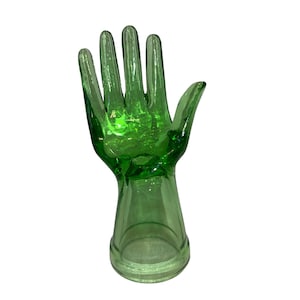 May include: A green glass hand sculpture with a base. The hand is open, with fingers extended. The glass is transparent, allowing light to pass through. The sculpture is a decorative item.