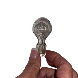 May include: A clear glass bottle stopper with a bulbous top and a detailed, textured design. The stopper has a short, cylindrical base and is held against a white background.