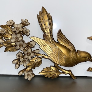 Pode incluir: Arte de parede de p&aacute;ssaros em metal dourado. Um p&aacute;ssaro em voo, com as asas abertas, pousado em um galho com flores brancas e douradas. O outro p&aacute;ssaro est&aacute; pousado em um galho. Decora&ccedil;&atilde;o para casa.