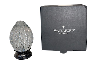 May include: A clear crystal egg-shaped decorative object with a faceted design, resting on a silver-colored base. The object is next to a dark gray box with the "Waterford Crystal" logo in white.
