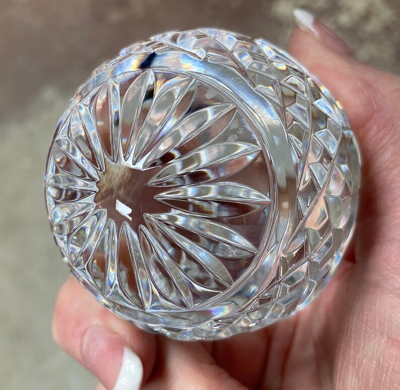 K&ouml;nnte beinhalten: Eine klare, geschliffene Glaskugel mit einem detaillierten Muster. Das Design zeigt ein zentrales Sonnenstrahlenmuster, umgeben von Diamanten und geometrischen Formen. Die Kugel wird in einer Hand gehalten, was die aufwendige Handwerkskunst und Lichtbrechung hervorhebt.