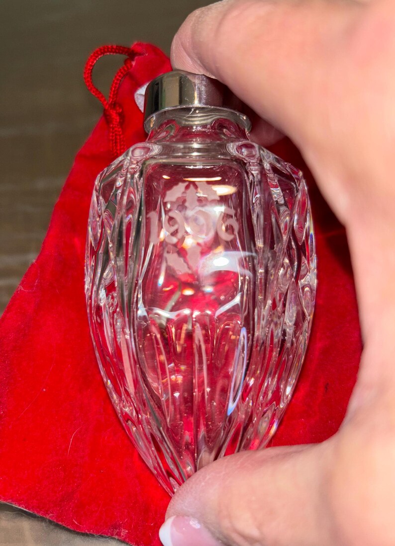 May include: A clear glass ornament with a silver top and the year 1996 etched on the front. The ornament is sitting on a red velvet bag.