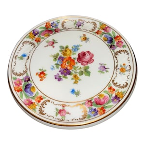 May include: A round white porcelain plate featuring a central bouquet of colorful flowers including red, orange, yellow, and purple blossoms. The rim is decorated with a floral garland and gold scrollwork accents on a white background.