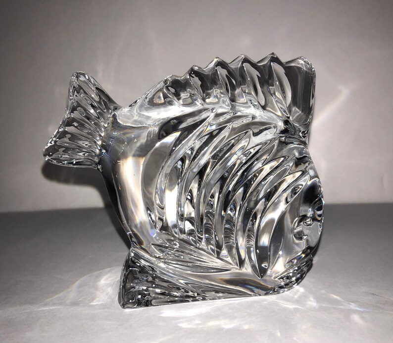 Peut inclure: Une sculpture de poisson en cristal transparent avec un design textur&eacute; d&eacute;taill&eacute;. Le poisson est tourn&eacute; vers la droite et a une nageoire sur le dessus de la t&ecirc;te.