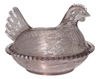 May include: A clear, light pink pressed glass hen on a nest dish. The lid features a detailed chicken figure with textured feathers, while the base is a round bowl with a beaded rim and a woven basket pattern embossed on the exterior surface.