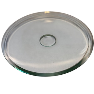 Puede incluir: Un plato de vidrio transparente y circular con un borde elevado y un pequeño agujero central. El plato es transparente, permitiendo el paso de la luz, y probablemente se utiliza para servir o exhibir. El diseño es simple y funcional.