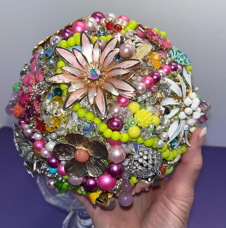 May include: A colorful bouquet made of vintage jewelry, beads, and brooches. The bouquet is round and features a pink flower with a rhinestone center, surrounded by green, yellow, and pink beads and brooches.