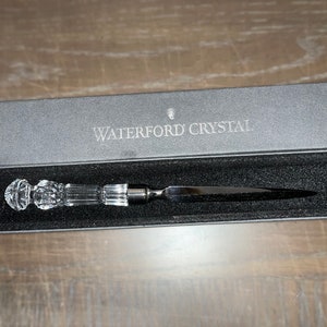 May include: A Waterford Crystal letter opener in a gray box. The opener has a clear crystal handle and a silver-colored blade. The box is open, revealing the opener nestled in black velvet. The words "WATERFORD CRYSTAL" are printed on the box.