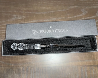 May include: A Waterford Crystal letter opener in a gray box. The opener has a clear crystal handle and a silver-colored blade. The box is open, revealing the opener nestled in black velvet. The words "WATERFORD CRYSTAL" are printed on the box.